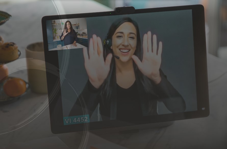 The image focuses on a tablet with an interpreter, wearing black clothes with a black background, signing and smiling. At the top left of the tablet is a woman, smiling and signing, in a kitchen. Behind the tablet, on a counter, are muffins and oranges.