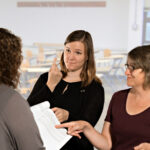 In a classroom, a group of women are communicating. Two women stand next to each other, one an interpreter, signing to a woman in front of them. The other woman is smiling and pointing at a paper in the woman’s hands.