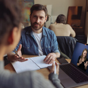 A man, wearing a blue shirt and white tee, is sitting in a coffee shop with a journal in front of him. He’s speaking to a woman, with a laptop between them on the table. On the laptop screen is an interpreter, signing in ASL.