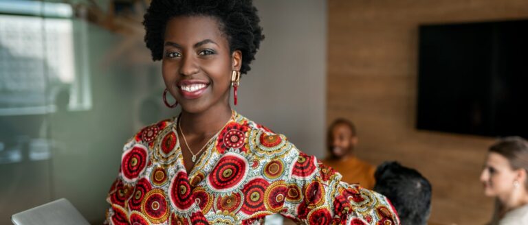 A Black woman with short black hair, wearing a patterned blouse and red earrings, is smiling. Behind her is a conference room with people sitting around a desk.