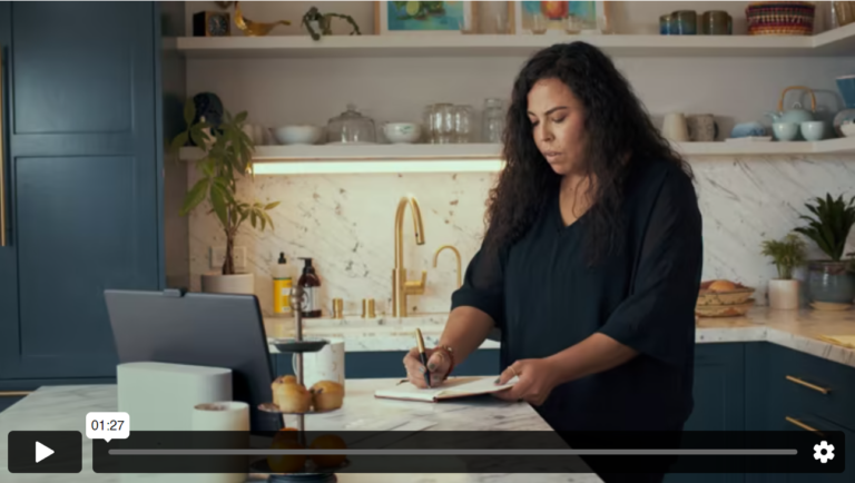 A woman with black hair and navy blouse is standing in a kitchen, writing in a journal. In front of her is a tablet.