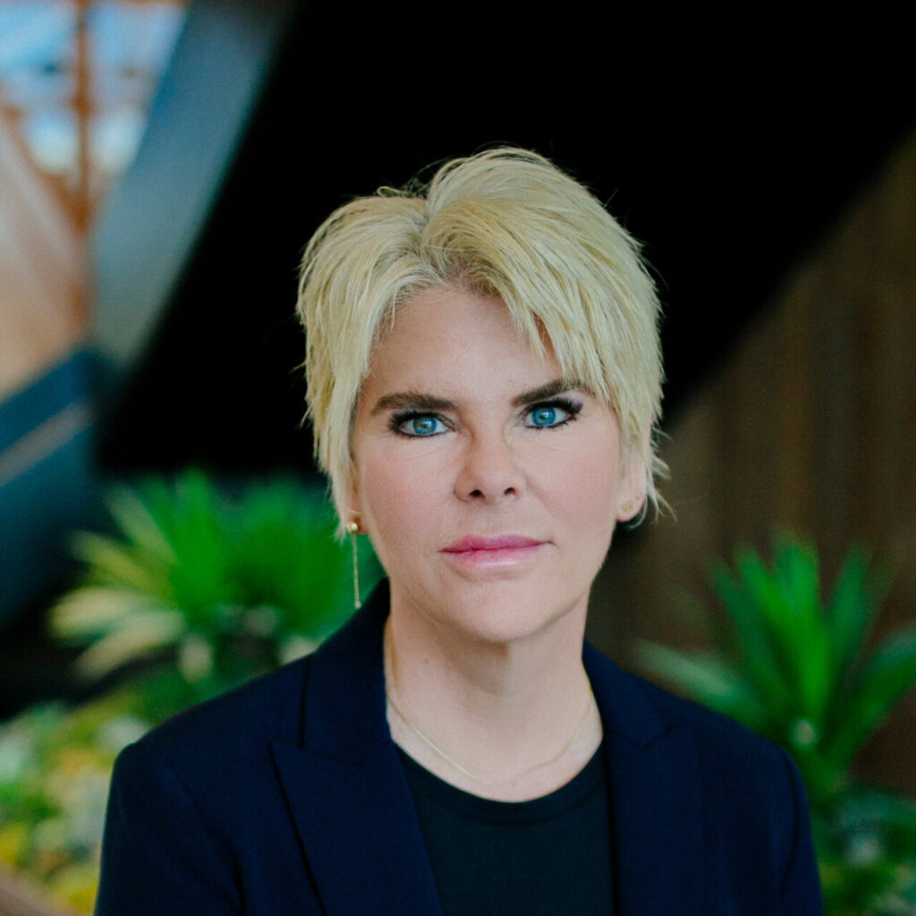 A white woman, ZP CEO Sherri Turpin, with short blonde hair, wearing a navy blazer and black shirt, is smiling and looking at the camera. Behind her is a brown and black wall surrounded by plants.