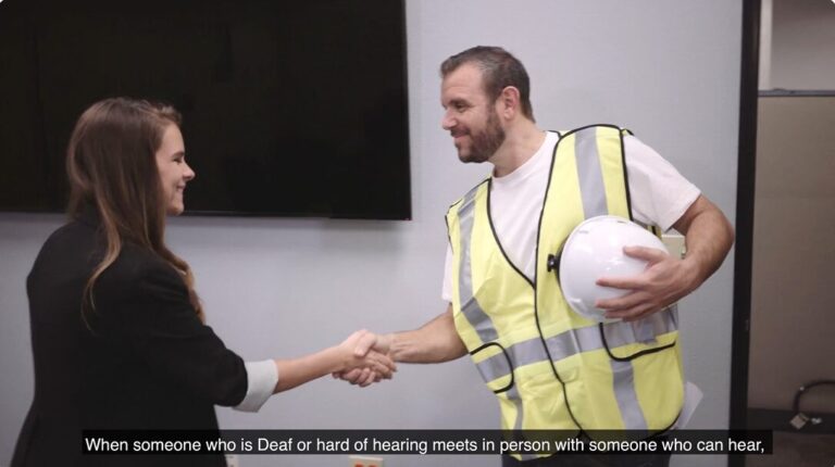 A woman in a blazer is smiling and shaking hands with a man wearing a safety vest and construction helmet.