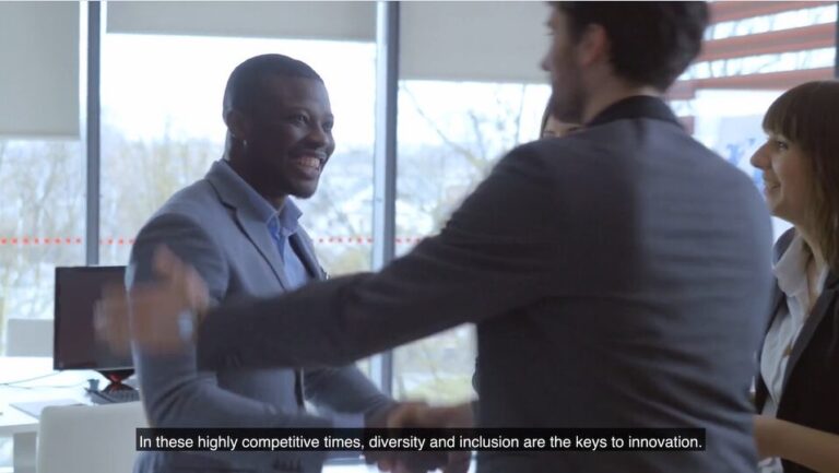 A video thumbnail of three people in an office. On the left is a man in a suit, smiling and shaking hands excitedly with the man in front of him. To the right is a woman, looking at the man and smiling.