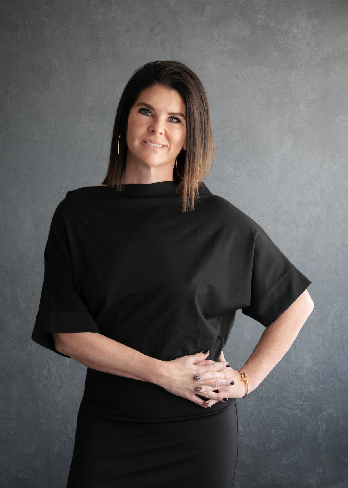 A woman, ZP CEO Sherri Turpin, with short brown hair is smiling and standing in front of a black background, wearing a black skirt and top, with her hands clasped on her hip.