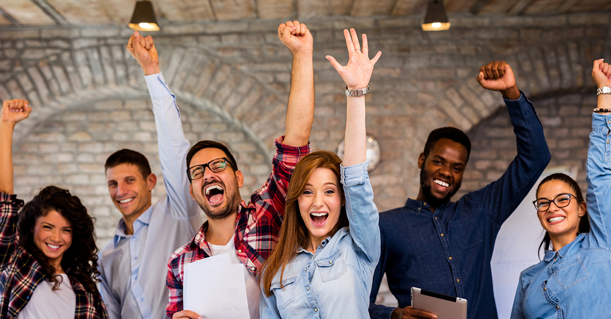 The image shows a group of people smiling and joyously raising their arms, standing in an office setting.