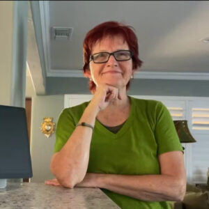 A white woman with ginger hair, wearing glasses and a green v-neck tee, is leaning on a counter in a home, looking ahead and smiling.