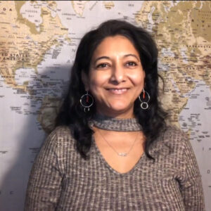 A woman with black hair, wearing a grey sweater and dangling earrings, is smiling. Behind her is a world map.