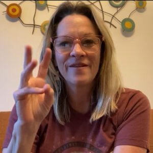 A white woman with blonde hair, wearing a red graphic tee and glasses, is signing and smiling.