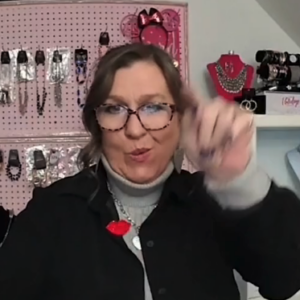 A woman with brown hair, wearing glasses, a grey turtleneck and black jacket, is signing in ASL. Behind her are many jewelry pieces on a pink wall.