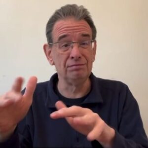 A white man with grey hair and glasses, wearing a navy collared long sleeve top, is signing in ASL.